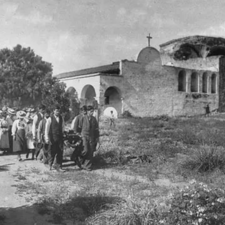 Historic procession of people near an old stone building with arches and a cross.