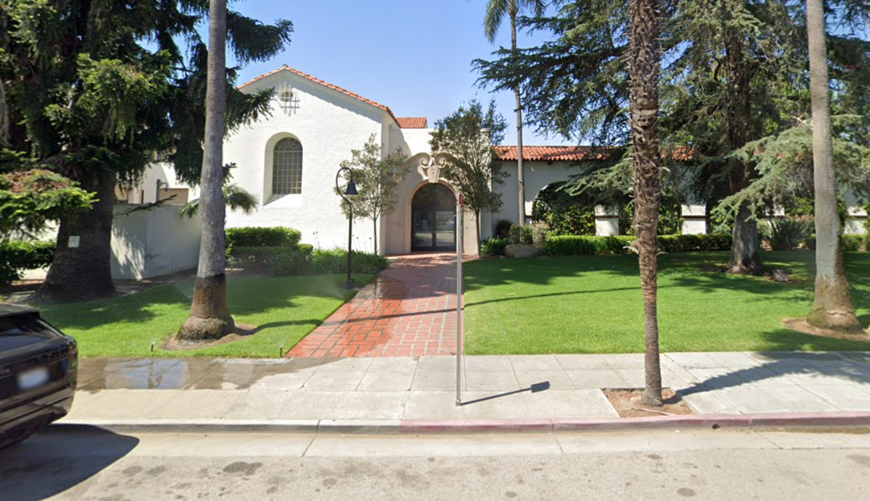 White building with red-tiled roof surrounded by trees and grass, viewed from the street.