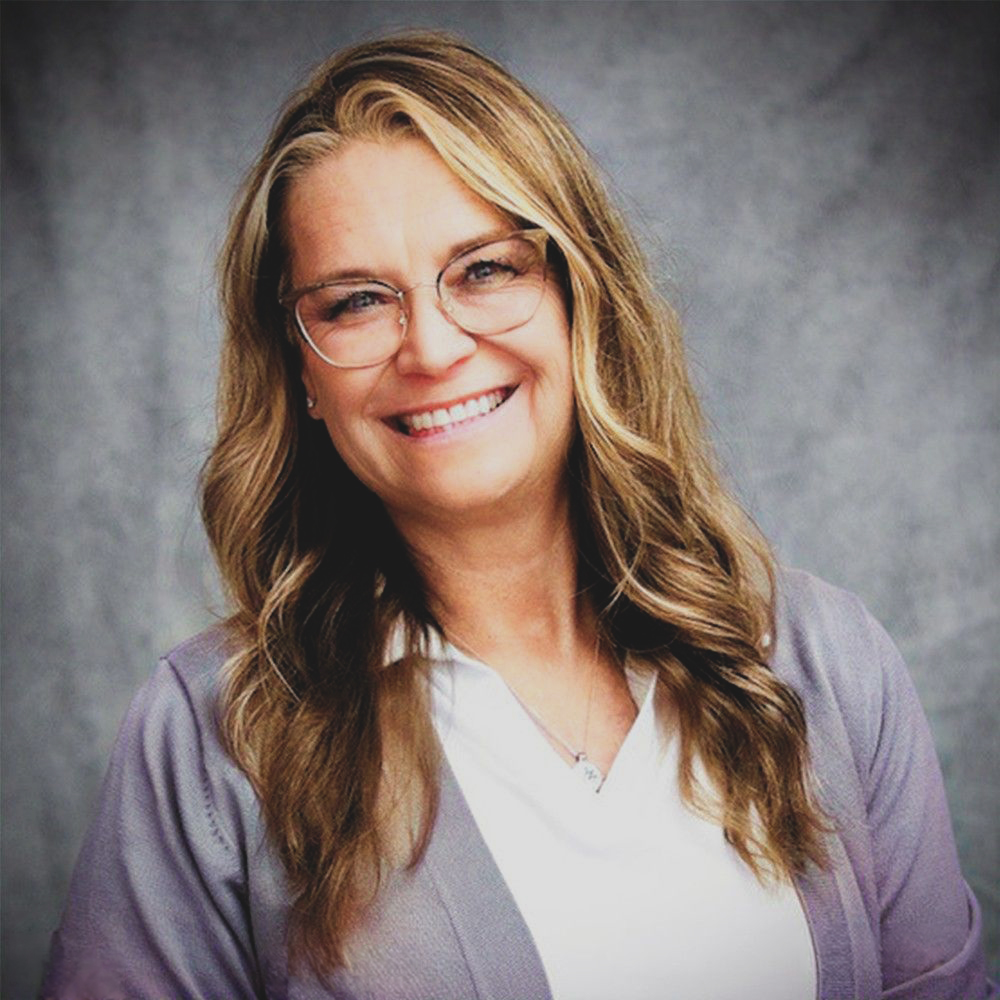 Woman with long hair, wearing glasses and a purple cardigan, smiling against a gray background.
