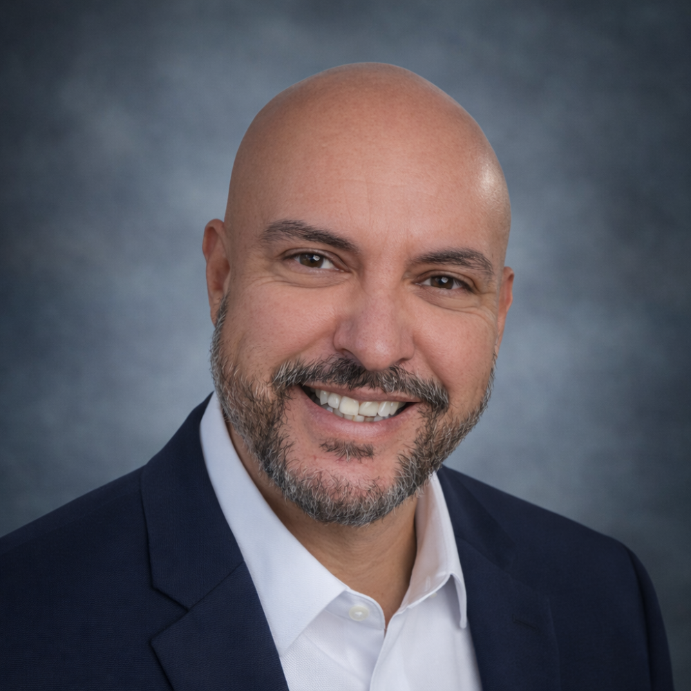 Smiling person with bald head and beard, wearing a dark suit and white shirt with a gray backdrop.