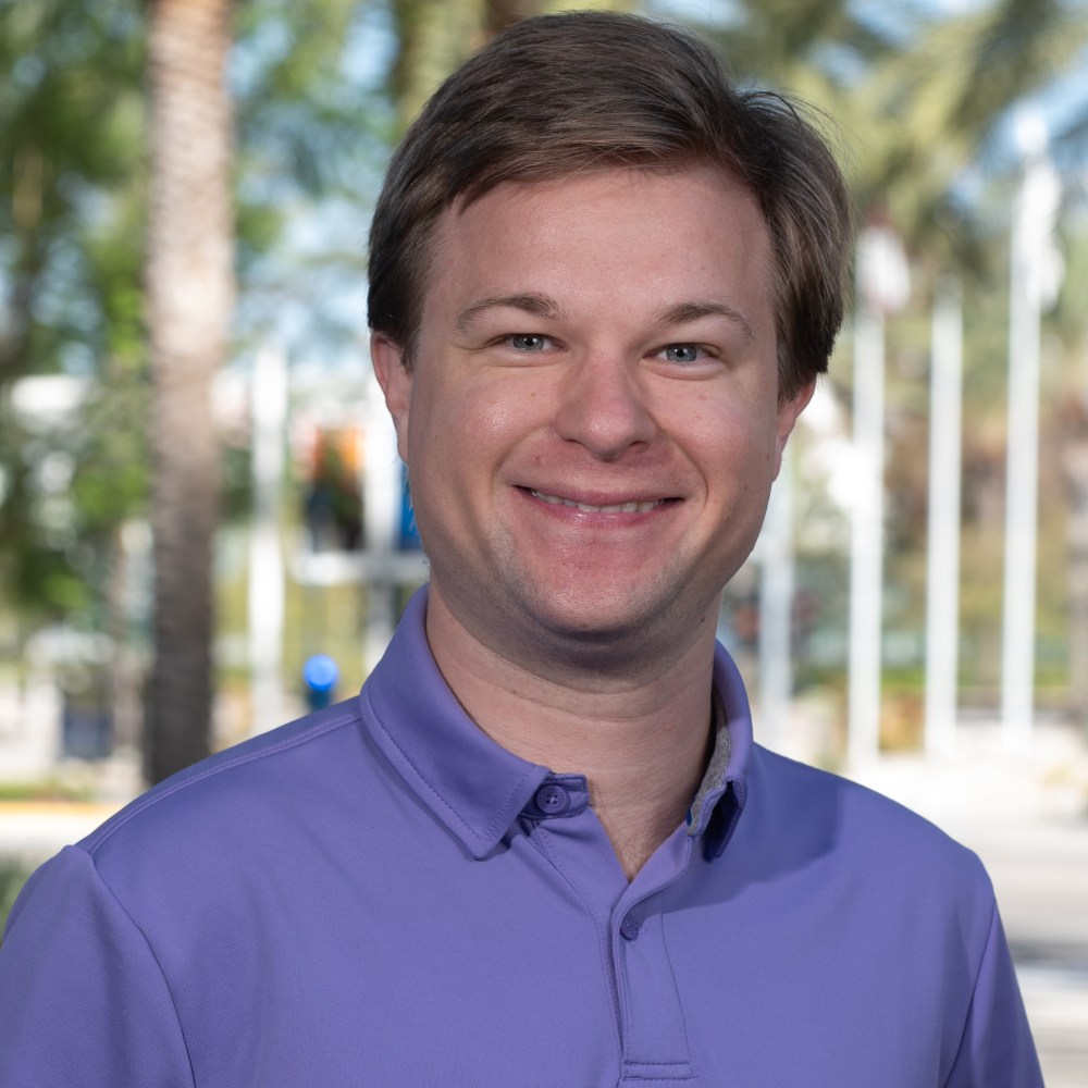 Man in a purple shirt smiling with trees in the background.
